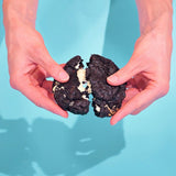 Cookies & Cream New York Cookie being broken in half showing black cocoa dough and white chocolate ganache filling by Crumbs & Doilies.
